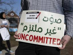 A sign in the foreground says "UNCOMMITTED" in red with Arabic text above, and a small image of a ballot with "uncommitted" checked and Biden unchecked. In the background is a woman in a hijab.