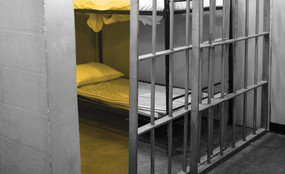 Photograph of an empty prison cell with the door open, showing an empty bed with no sheets.