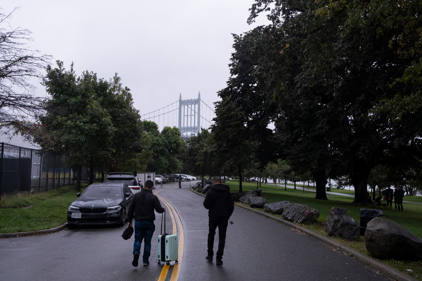 two men walk on the road rolling a suitcase with a bridge in the distance