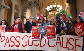 Activists and New York state Senator Julia Salazar rally to pass good cause eviction in Albany. They stand in the high-ceilinged halls of the Capitol with a white banner reading PASS GOOD CAUSE and various red signs.