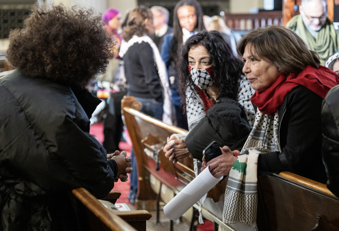 Attendees of the “Emergency Summit for Gaza” gathered at the Rainbow Push Coalition’s headquarters in Chicago. Two attendees right of center lean over a pew. One of the attendees is wearing a keffiyeh-patterned mask. The other attendee to their right is also wearing a keffiyeh. There is another attendee, leaning over their respective pew with their back turned towards the camera, in the left side of the image conversing with the other two attendees.