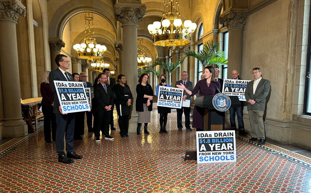 A group of people holds a press conference in Albany, New York, holding signs that read "IDAs DRAIN $1.8 BILLION A YEAR FROM NEW YORK SCHOOLS."