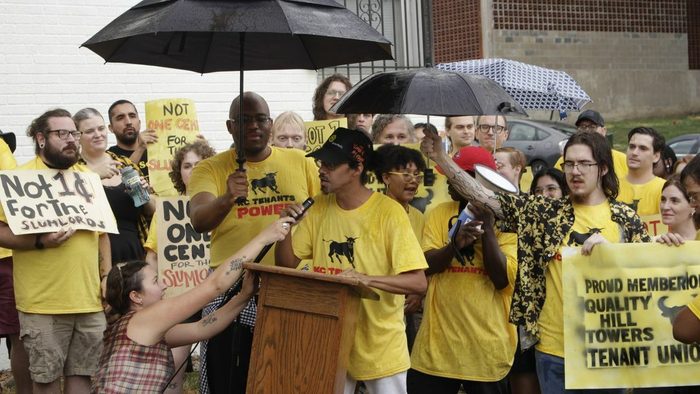 A crowd of people in yellow shirts gesture behind a podium.