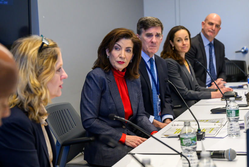 Governor Kathy Hochul, wearing a red turtleneck and blazer, sits at a table with other officials.
