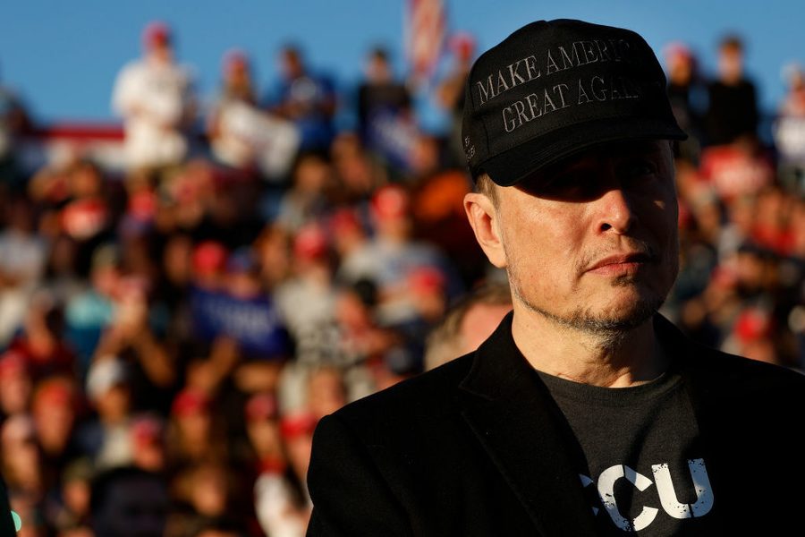 Elon Musk wears a MAGA hat, his eyes in shadow, with a blurry crowd behind him