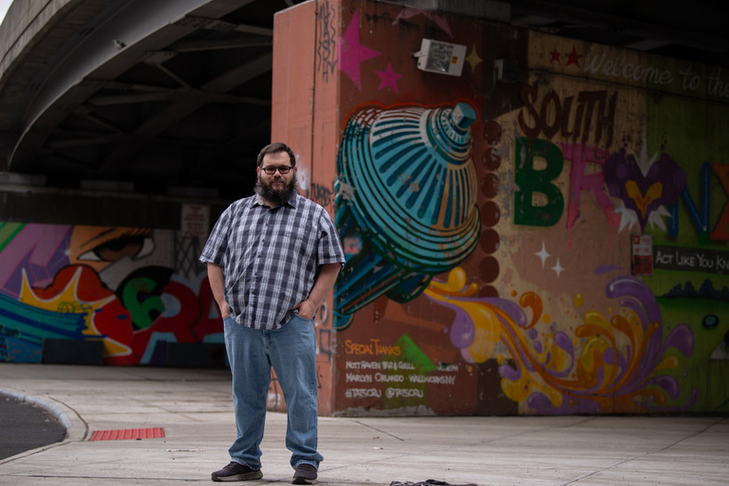 A man in a plaid short-sleeve shirt stands with his hands in his pockets under a painted bridge in the Bronx.