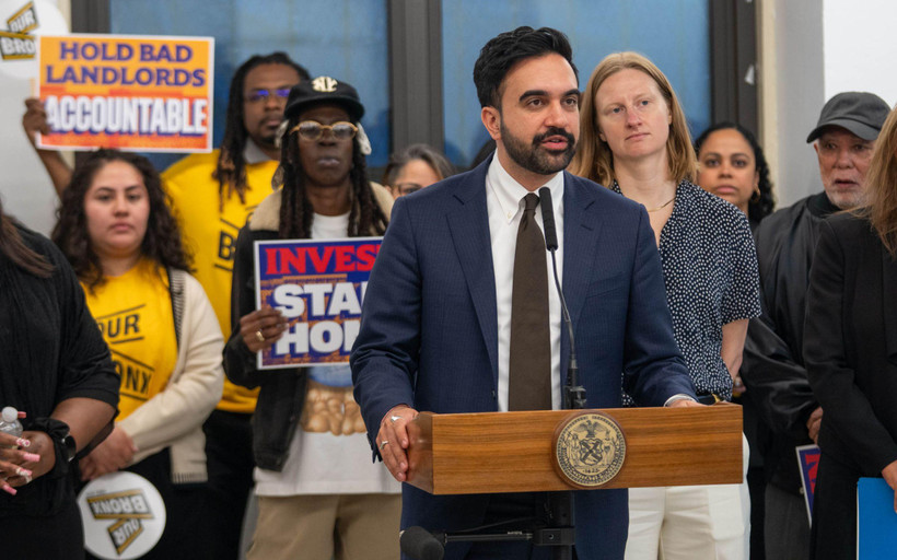At a press conference, Mayor Zohran Mamdani stands speaking at a podium, behind and to his left stands Cea Weaver, director of the New York City Mayor's Office to Protect Tenants. Behind them stand people holding signs bearing the words "Hold bad landlords accountable" and "Invest in stables homes."