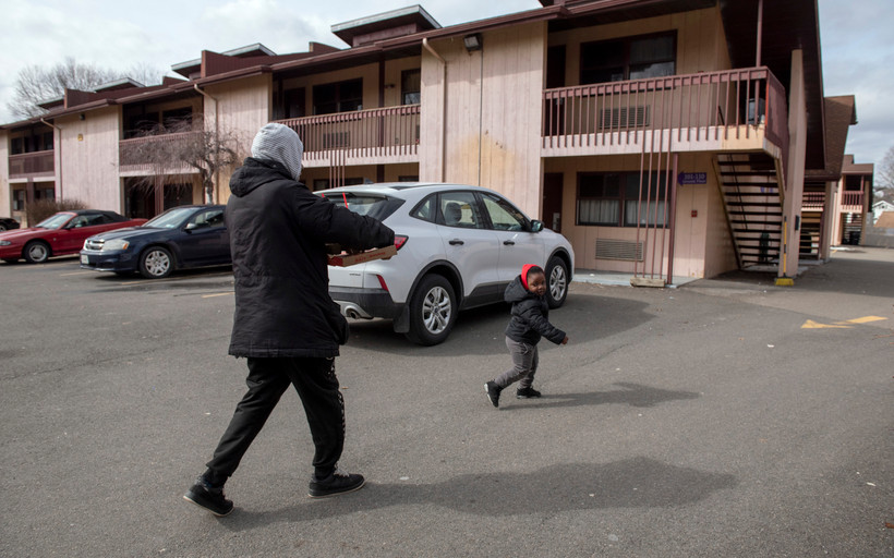 An adult and child wearing winter coats walk across a motel parking lot with three cars parked in it. The child is smiling and turns back to look at the adult, who is facing away from the camera.