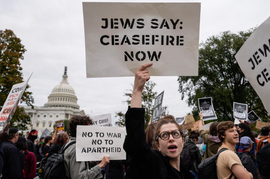 A person holds up a sign that reads "JEWS SAY: CEASEFIRE NOW". Behind them is another sign that reads "NO TO WAR NO TO APARTHEID". The U.S. Capitol Building is seen in the background, behind the crowd of protestors.