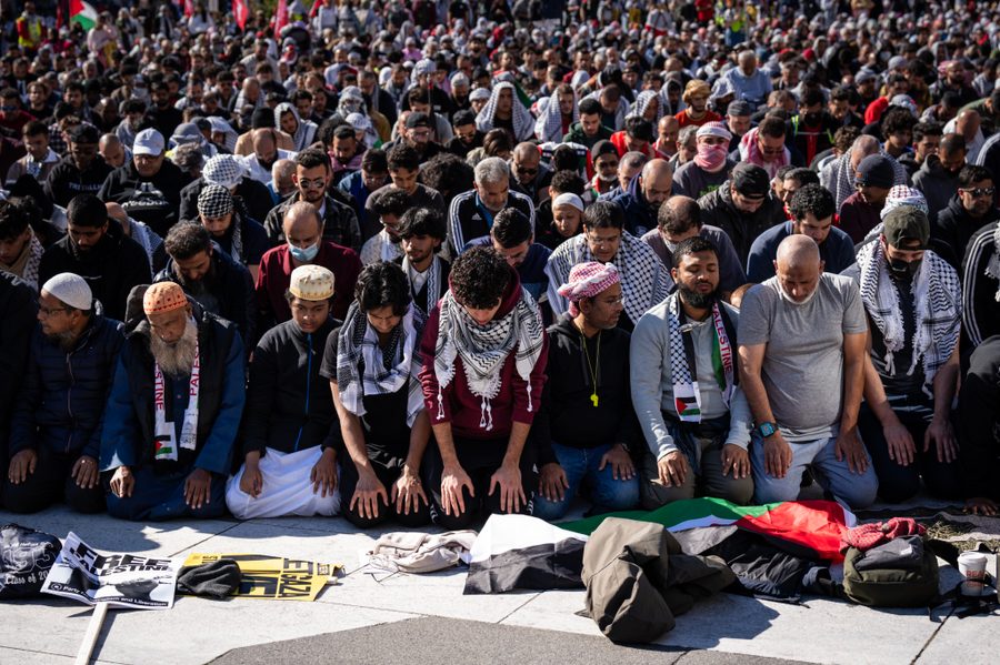 Hundreds of protestors kneel on the ground in prayer, and fill the image kneeling shoulder to shoulder. Some are wearing keffiyehs, and a Palestinian flag is on the ground at the forefront of the picture.