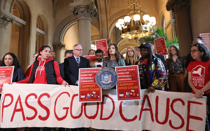 Activists and New York state Senator Julia Salazar rally to pass good cause eviction in Albany. They stand in the high-ceilinged halls of the Capitol with a white banner reading PASS GOOD CAUSE and various red signs.