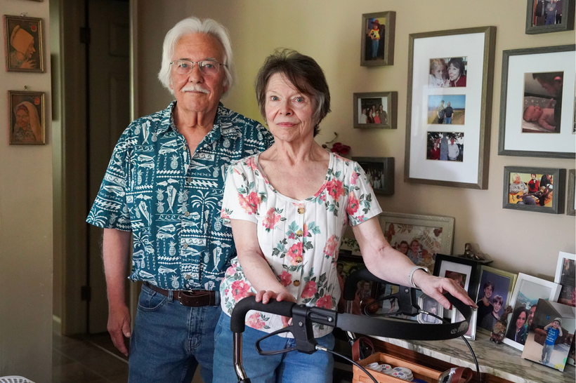 An older man and woman stand in a room filled with photographs from the past, looking at the camera. The woman holds a walker.