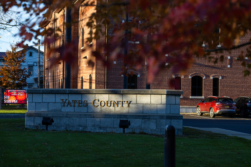 An image of the sign of a concrete sign for Yates County, partially lit by sunlight and partially clouded. Blurred fall leaves frame the image.