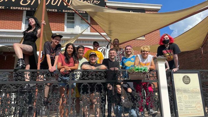 Grinning people in punk attire pose on a railing outside a coffee shop