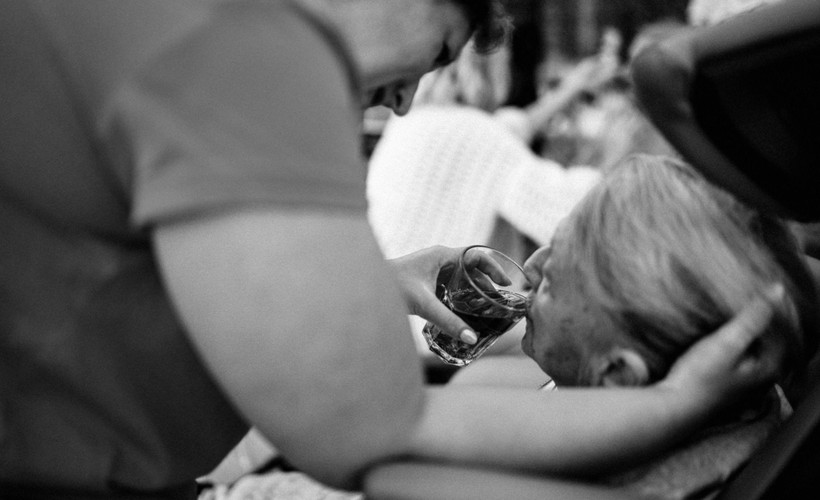 A standing woman wearing scrubs gently cradles a seated older woman's head while she helps her drink from a glass.