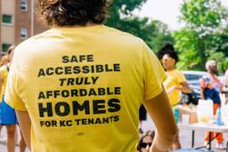 A masculine figure in a baseball cap stands with his back to the camera. His bright yellow shirt reads "Safe accessible truly affordable homes -- KC Tenants Union". Divers figures in yellow shirts stand int he background.