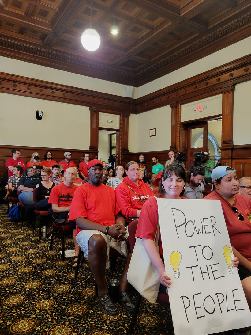 Activists dressed in red sit at the Rochester City Council meeting. A woman at the front holds a sign reading "power to the people."