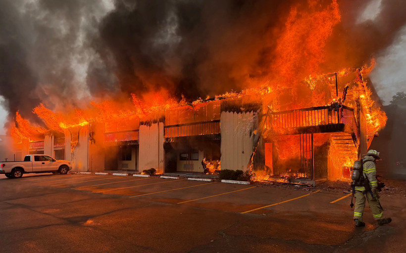 A motel is completely engulfed in flames, and black and gray smoke pours into the sky. A lone firefighter walks in the right side of the frame.