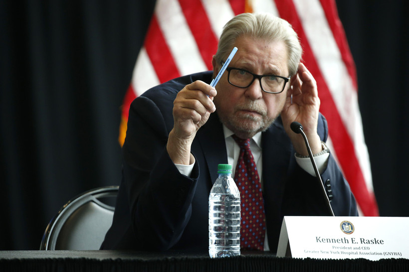 Greater New York Hospital Association President Kenneth Raske sits at a table and gestures with a pen in front of an American flag.