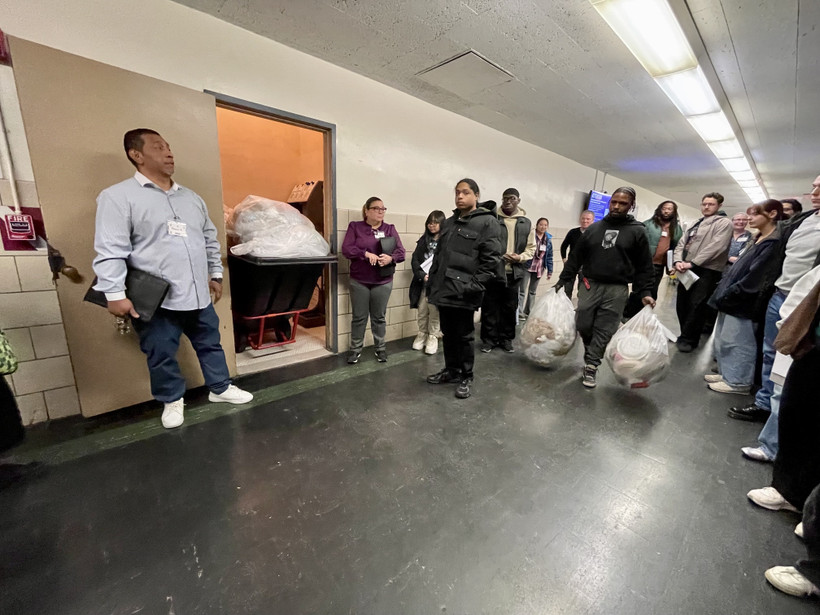 A group of people stand in a fluorescent-lit hallway. One man carries bags of trash through the group to a trash room that opens off the hallway.