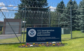 A blue sign reading "U.S. Immigration and Customs Enforcement, Buffalo Detention Center, Batavia New York" sits on the grass in front of a tall metal fence with barbed wire at the top.