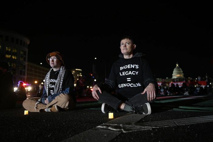 Two demonstrators sit crosslegged on a road at night with shirts reading "Biden's legacy = genocide." In the background is a crowd in front of the Capitol building.