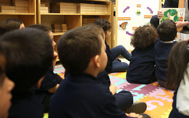 Preschool-aged children sit facing away from the camera on a colorful puzzle mat of numbers, while an adult at the front of the room shows them a page from The Very Hungry Caterpillar.