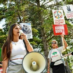 A woman protester shouts into a large megaphone in the foreground. A few older protesters stand behind her with signs reading "Gaza love," "Abandon Biden--ceasefire now" and have flower and peace sign imagery.