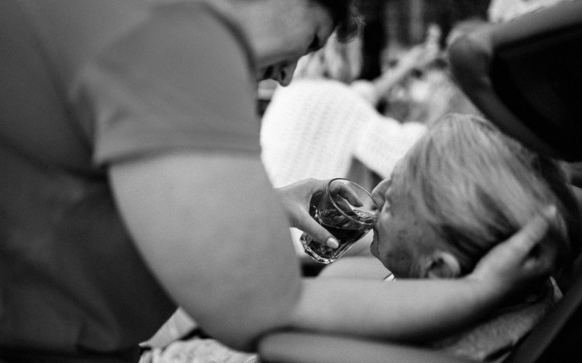 A standing woman wearing scrubs gently cradles a seated older woman's head while she helps her drink from a glass.