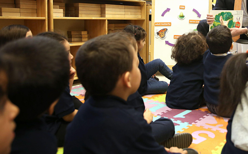 Preschool-aged children sit facing away from the camera on a colorful puzzle mat of numbers, while an adult at the front of the room shows them a page from The Very Hungry Caterpillar.
