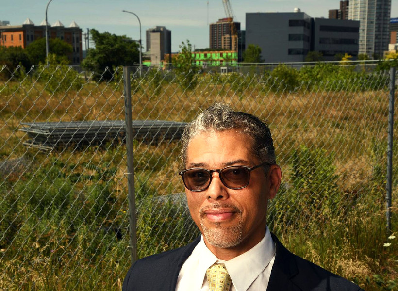 Charles Garland stands in front of a chain link fence with a field and buildings behind him.