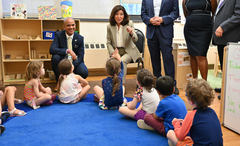 New York City Governor Kathy Hochul sits in front of a group of small children on a blue carpet.