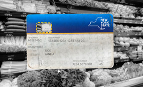 A New York state benefits card with a cut-out yellow outline where a digital chip would go. In the background, a black and white photo of produce in  a grocery store.