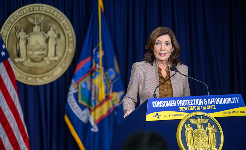 New York Governor Kathy Hochul stands at a blue and yellow podium that says "Consumer Protection & Affordability / 2024 State of the State."