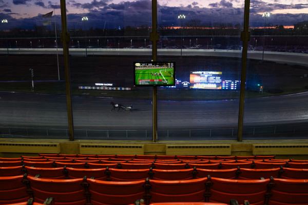 Rows of empty seats watch a horse racing track with a lone harness racer and horse.