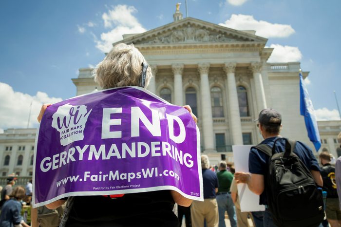 A person stands with their back facing the camera in front of the WI state capital. They are holding a purple sign on their back that says "END GERRYMANDERING" with the logo for WI Fair Maps Coalition and "www.FairMapsWI.com". Other people with signs can be seen in the background.