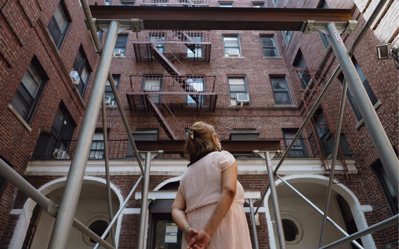 A woman stands outside a brick building, looking away from the camera, with her hands clasped behind her back.