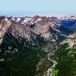 Photo of the Crazy Mountain Range in Montana. Mountains in the distance have snow on them in the background. In the foreground is a lush, green mountain valley.