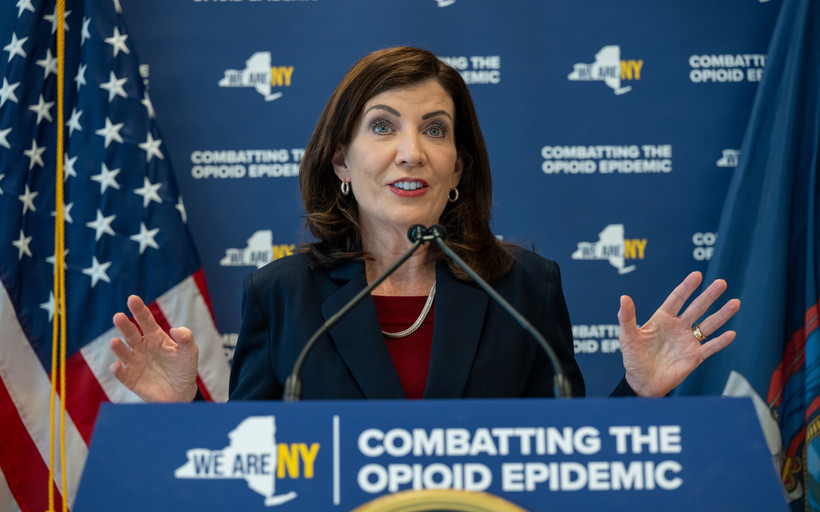 Kathy Hochul stands with her hands spread at a podium reading "Combatting the Opioid Epidemic"