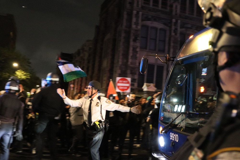 A police officer in a white shirt at night with a Palestinian flag flying behind him and an MTA bus on the right side of the frame