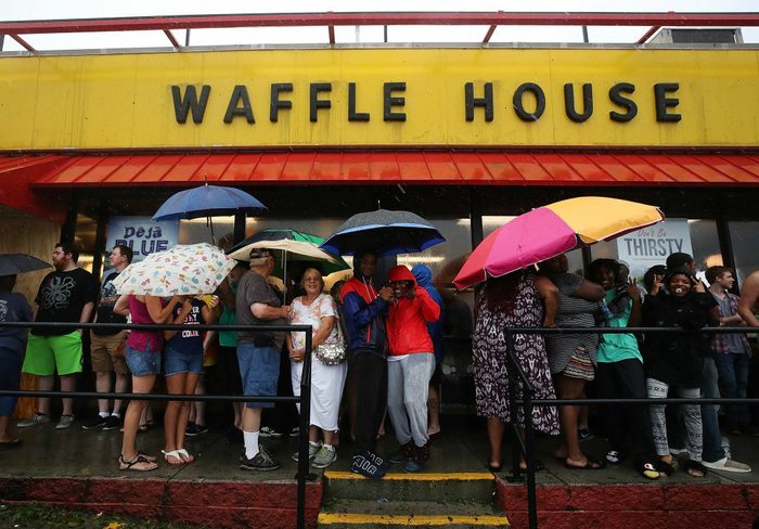 Below a yellow Waffle House awning, a crowd of people with umbrellas