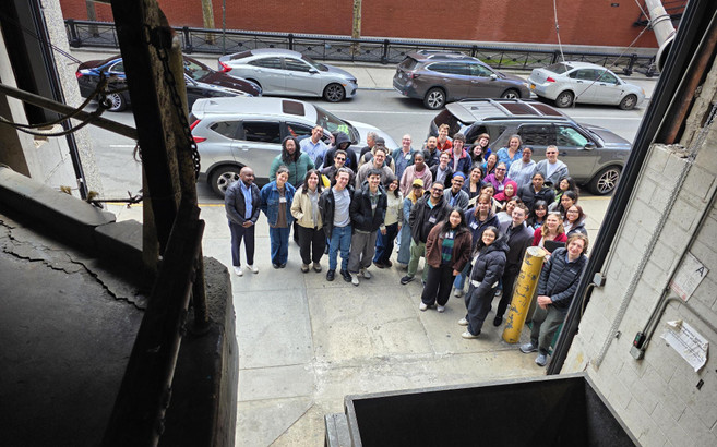 A group of about 50 people pose for a photo taken from inside a garage housing a trash compactor.