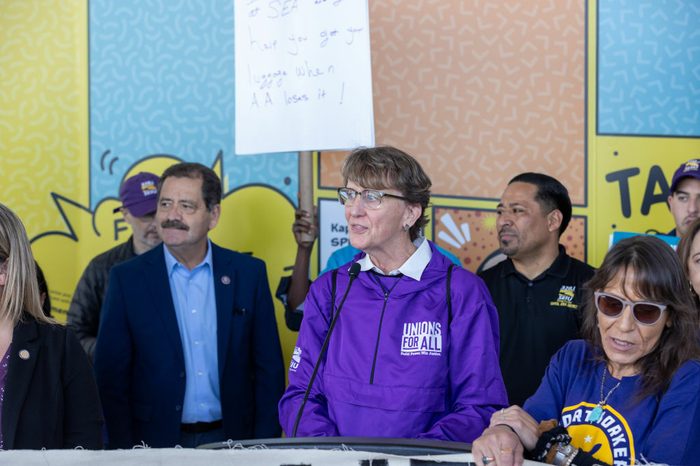 SEIU president Mary Kay Henry and Congressman Jesús “Chuy” García stand in front of a rally surrounded by SEIU members. Henry is wearing a purple SEIU Unions for All windbreaker speaks into a microphone in the center right of the image. García stands to her left wearing an SEIU pin.