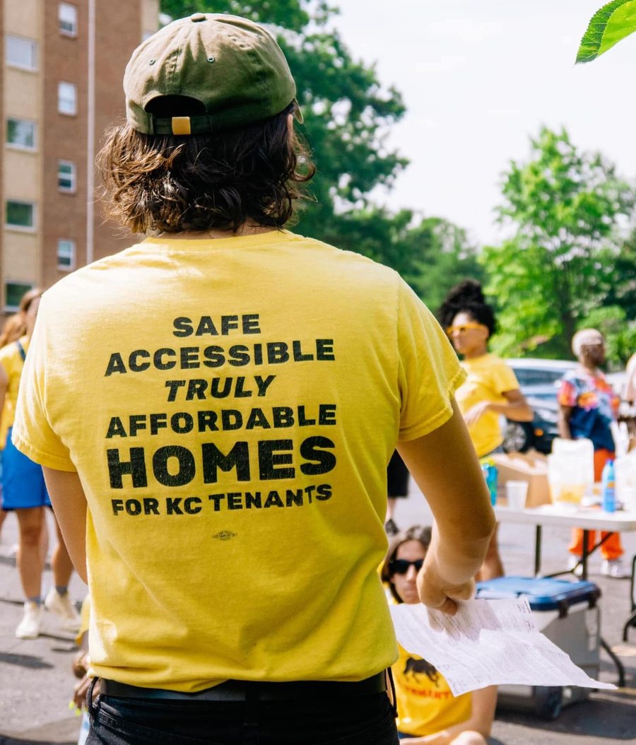 A masculine figure in a baseball cap stands with his back to the camera. His bright yellow shirt reads "Safe accessible truly affordable homes -- KC Tenants Union". Divers figures in yellow shirts stand int he background.