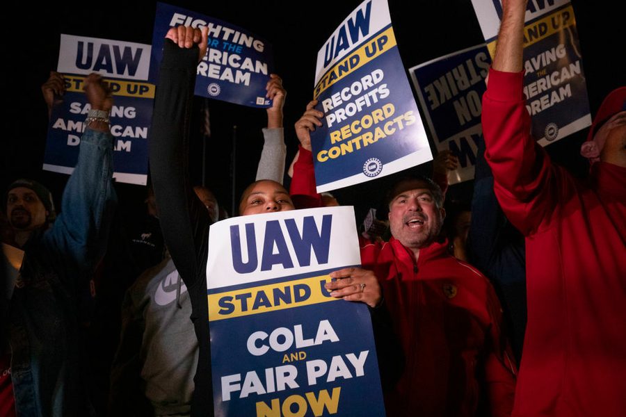 A handful of cheering autoworkers hold signs reading "UAW Stand Up COLA Fair Pay Now" and "UAW Stand Up Record Profits Record Contracts."