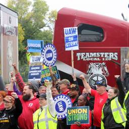 A crowd of striking workers hold up UAW signs and fists, standing in front of two trucks that say Teamsters.
