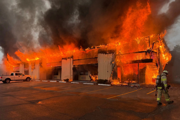 A motel is completely engulfed in flames, and black and gray smoke pours into the sky. A lone firefighter walks in the right side of the frame.