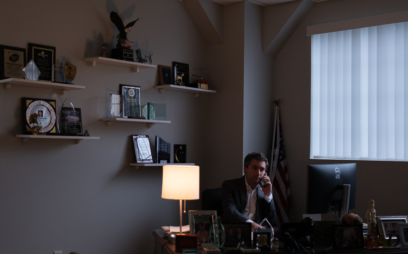 Senator James Skoufis sits at a large, dark desk illuminated by the soft glow of a lamp, speaking on the phone and looking away from the camera. Plaques and awards adorn the wall and desk.