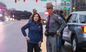 Governor Kathy Hochul stands with a hand on her hip next to a New York state trooper on a cloudy day.