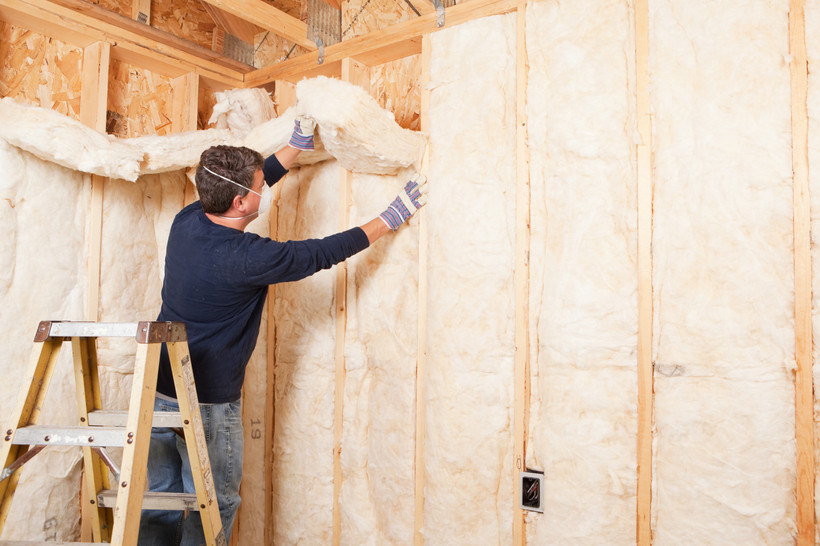 A man wearing a mask installs pink fiberglass insulation on the inside of a home.